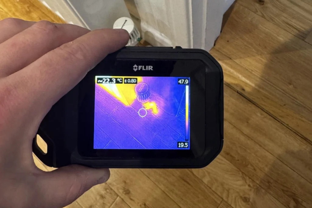 FLIR thermal imaging camera checking heat levels around a radiator valve and wooden floor.