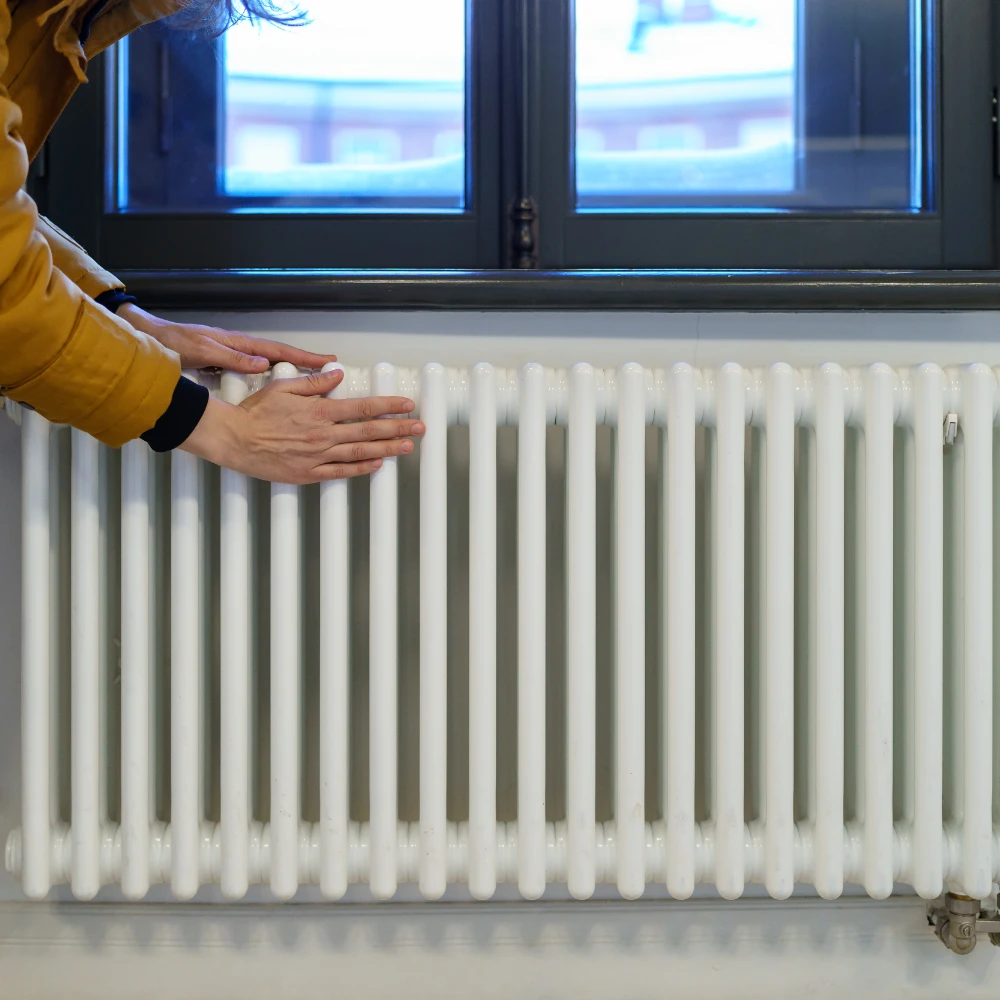 Woman placing her hands on a white column radiator beneath a window to check its warmth.