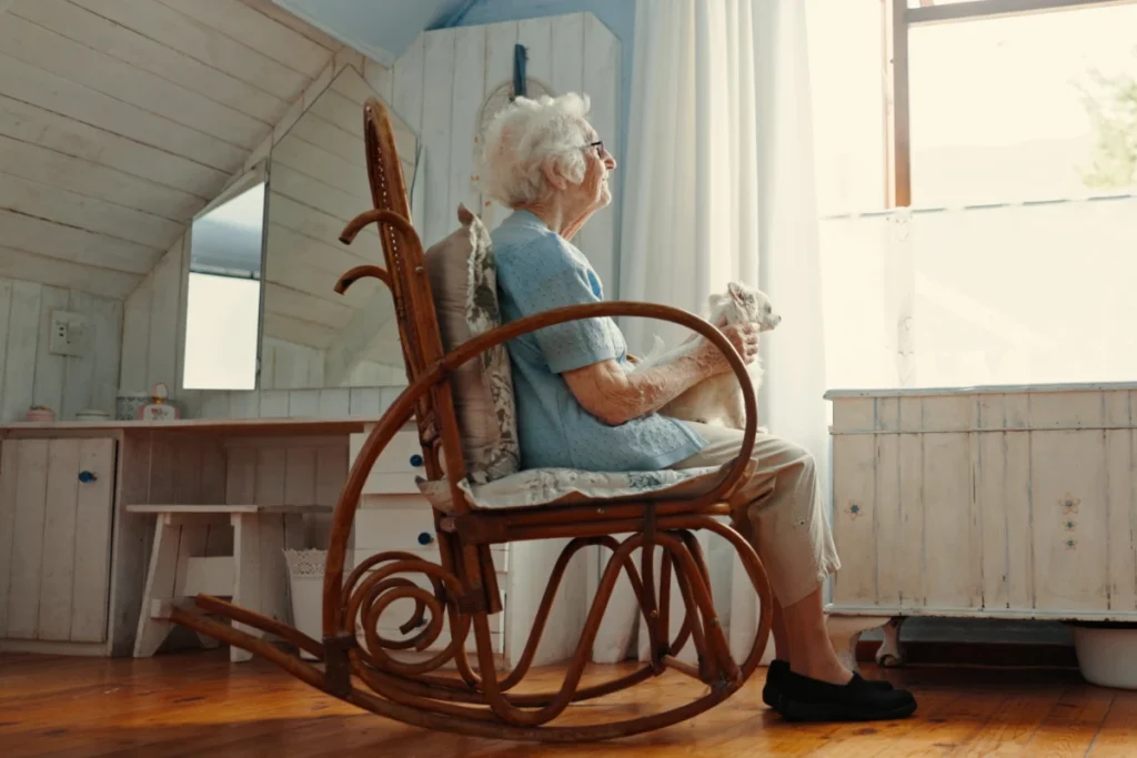 Elderly woman relaxing in a wooden rocking chair with a small white dog beside a sunlit window.