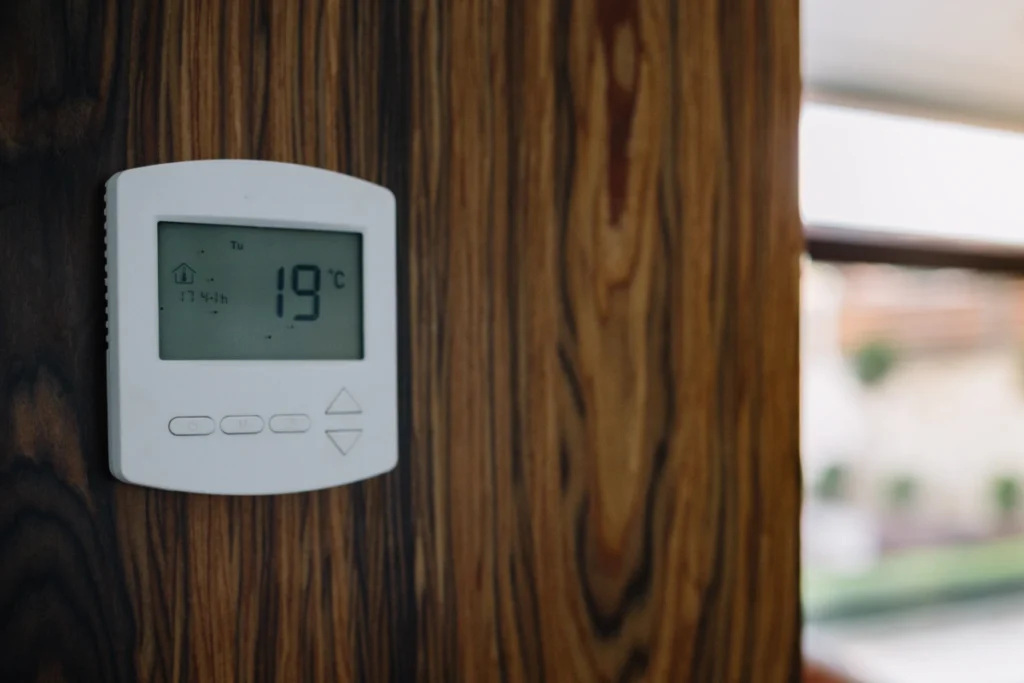White digital room thermostat displaying 19°C, mounted on a dark wooden wall beside a window.