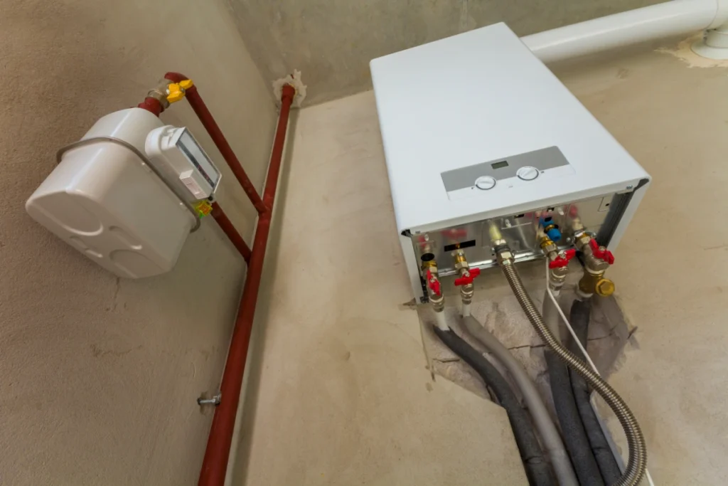 White wall-mounted gas boiler with exposed pipework, isolation valves and a gas meter.