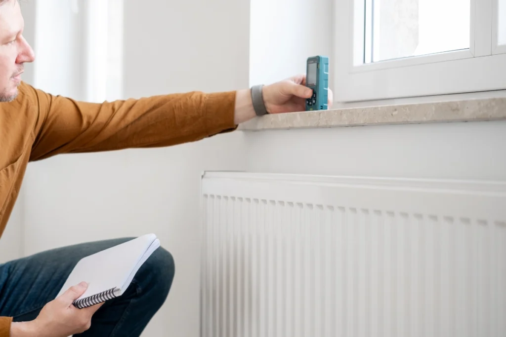 Heating engineer using a digital measuring device beside a radiator and window in a residential property.