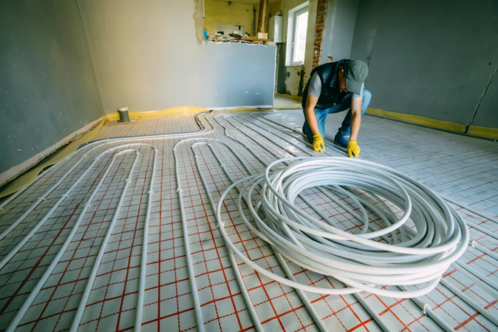 Heating engineer installing white underfloor heating pipes across insulation panels during a residential renovation.