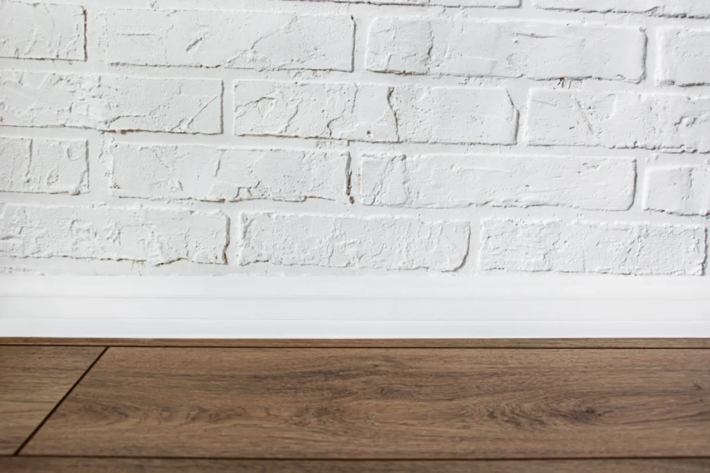 White skirting board fitted beneath a painted brick wall above dark wooden flooring.