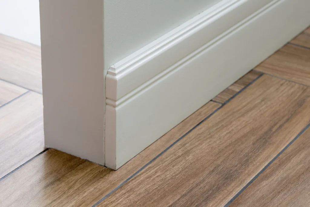 Close-up of white skirting boards fitted along a wall above wood-effect floor tiles